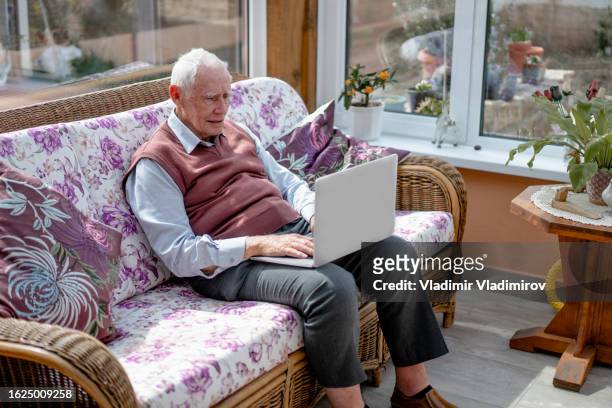 um idoso está sentado em casa, em um sofá com um laptop no joelho enquanto navega na rede - lockdown - fotografias e filmes do acervo