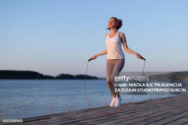 beautiful young girl in her 20s with blonde hair by the river on a fitness path exercising with jumping rope while listening to music and enjoying the sunset - woman skipping stock pictures, royalty-free photos & images