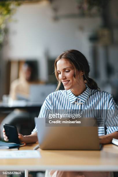 a happy beautiful blonde businesswoman holding her mobile phone while working on her computer - marketing expertise stock pictures, royalty-free photos & images