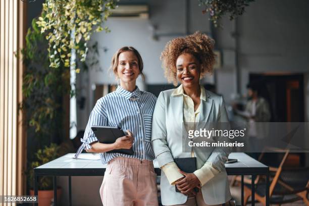 two happy beautiful blonde businesswomen looking at camera while standing in the office - two people portrait stock pictures, royalty-free photos & images