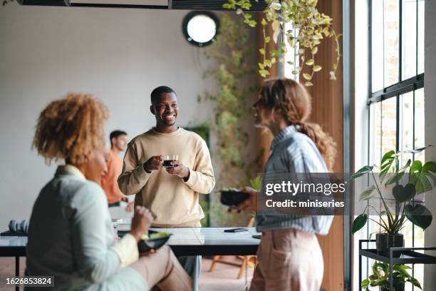 a happy businessman with a cup of tea talking with his coworkers while standing in the office - business people group brown stock pictures, royalty-free photos & images