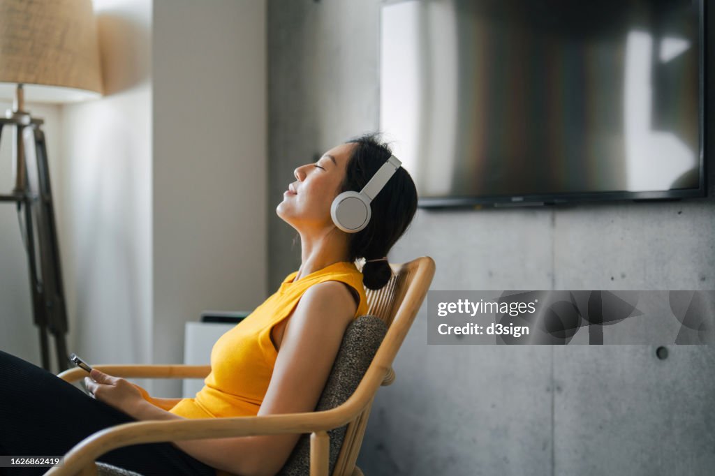 Beautiful young Asian woman with eyes closed enjoying music over headphones from her smartphone while relaxing on rattan arm chair at cozy home. Music, lifestyle and technology