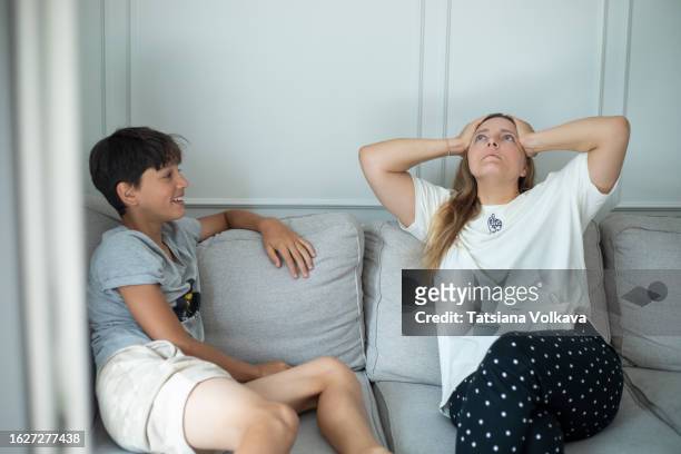 numb teenager sitting on sofa and looking at lost mom who keeping hands on head and looking up with amazed eyes, with fake smile full of despair throughout talk of adolescence depression. - fake smile stock-fotos und bilder