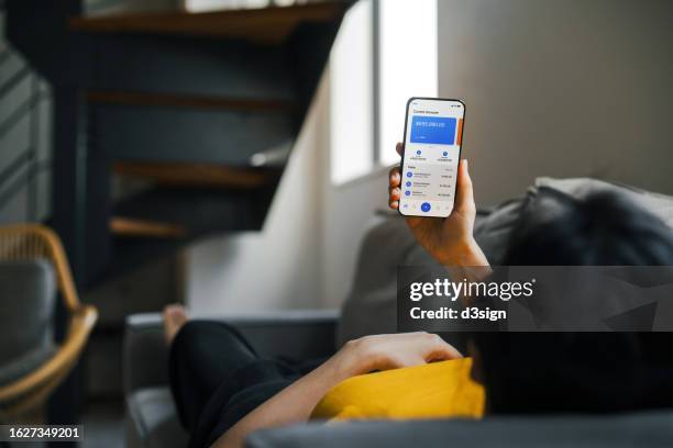 young asian woman using smartphone while lying on the sofa at cozy home, checking her bank account via online banking and managing her finances. online personal banking. wealth management. digital banking habits. smart banking with technology - online bank statement stock pictures, royalty-free photos & images