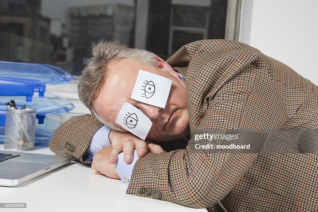 Businessman sleeping with sticky notes on eyes at desk in office