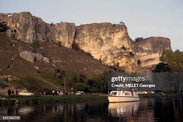 River Tourism. En France, tourisme fluvial, en 1971 : un bateau de plaisance piloté par un homme avec une casquette de marin, passant sur une rivière...