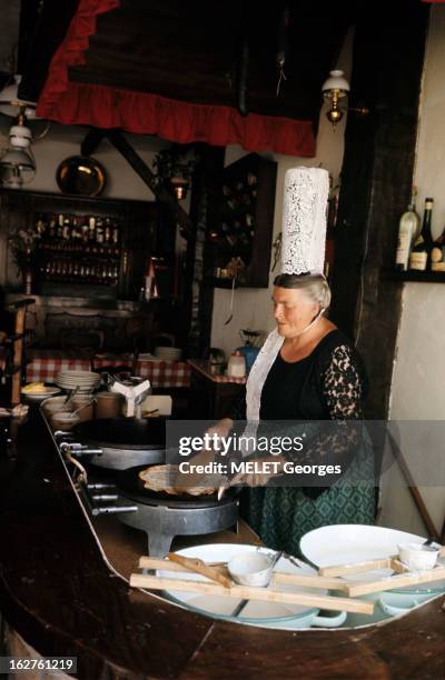 Brittany. En 1971, en Bretagne, dans une cuisine, une bretonne en tenue traditionnelle, avec une coiffe, faisant des crêpes.