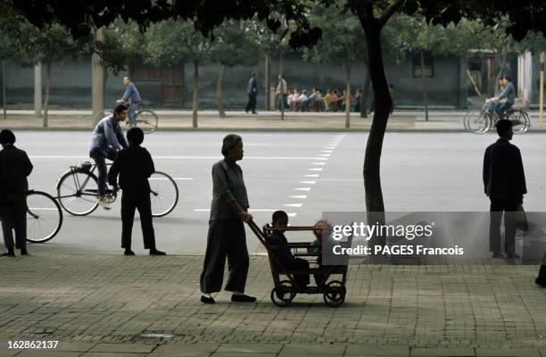 China. En Chine, dans une rue, où circulent quelques cyclistes, une femme, en pantalon avec une veste chinoise et des ballerines, poussant sur le...