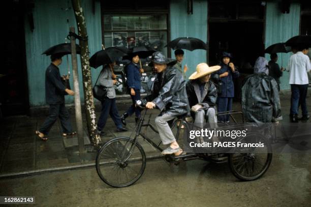 China. En Chine, à Shangaï, dans une rue un jour de pluie, des passants avec des parapluies et un homme, avec un imperméable et un chapeau de pluie,...