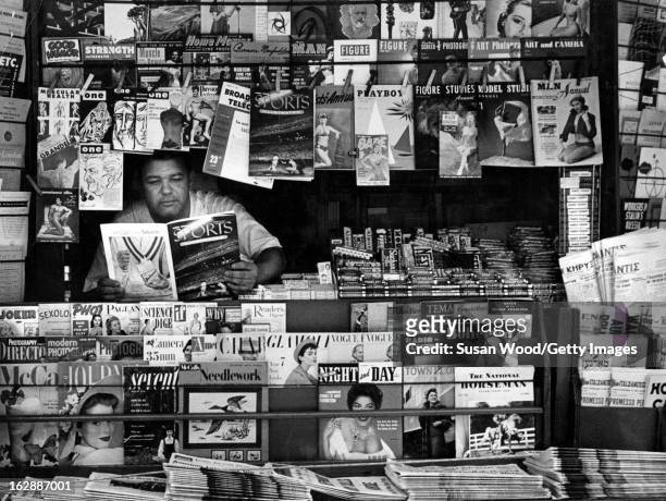 At a newstand, a vendor reads an issue of Sports Illustrated magazine as he waits for cistomers, New York, New York, August 1954.