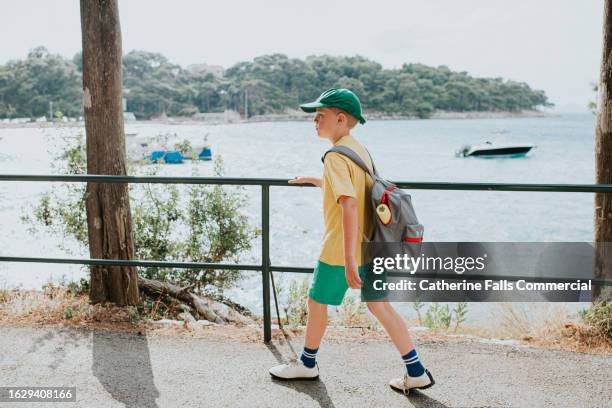 a little boy walks along a road, beside the sea, wearing casual clothing and carrying a backpack - one embankment stock pictures, royalty-free photos & images