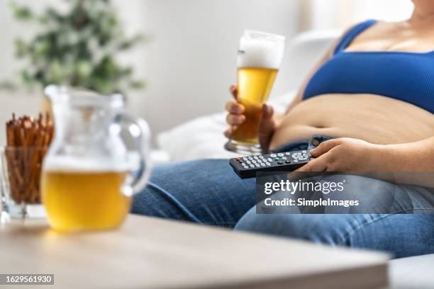 an obese woman overeats by eating junk food, drinking beer and watching tv. - obesity imagens e fotografias de stock