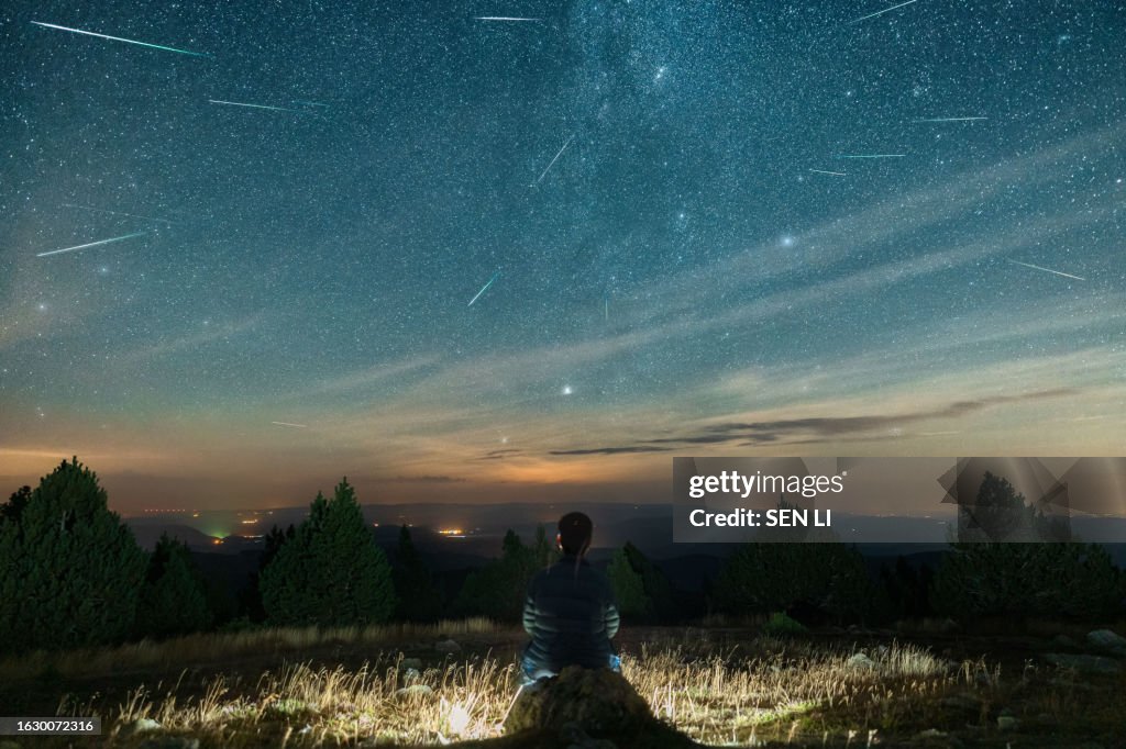 A man sits on top of a mountain watching the Perseid meteor shower