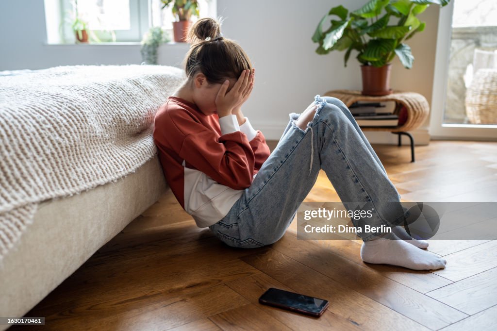 Unhappy teen girl covering face with hands crying while sitting on floor with mobile phone nearby