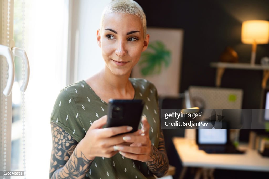 Woman with short hair and sleeve tattoo looking through window while using mobile phone