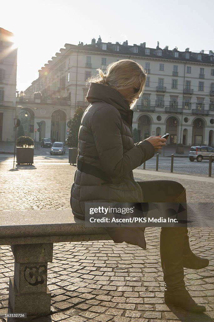 Woman sends text from bench in piazza, sunrise