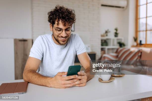 young smiling casually clothed man text messaging on smart phone at home - positivo imagens e fotografias de stock