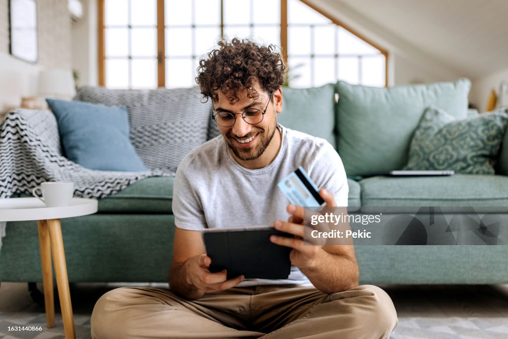 Junger lässig gekleideter Mann, der online einkauft, während er zu Hause auf dem Boden sitzt