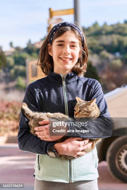 a girl smiling, carrying a cat, looking at the camera, in the background you can see part of a vehicle, trees and a blurred sign. - cat holding sign stock pictures, royalty-free photos & images