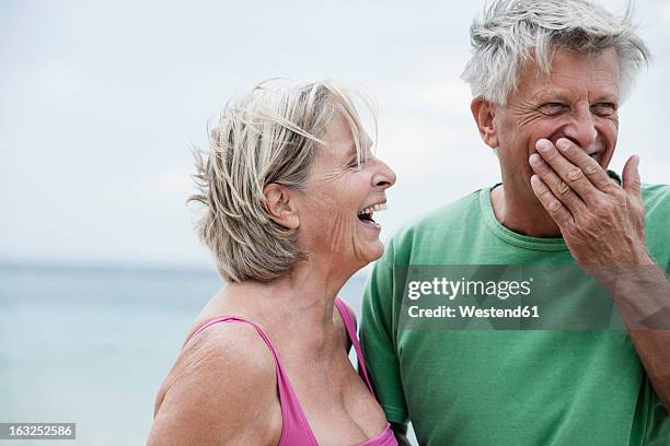 spain, senior couple smiling on beach - mano tapando la boca fotografías e imágenes de stock