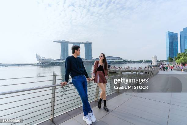 tourist couple walking along marina bay with merlion park and buildings in the background. - singapore stock pictures, royalty-free photos & images