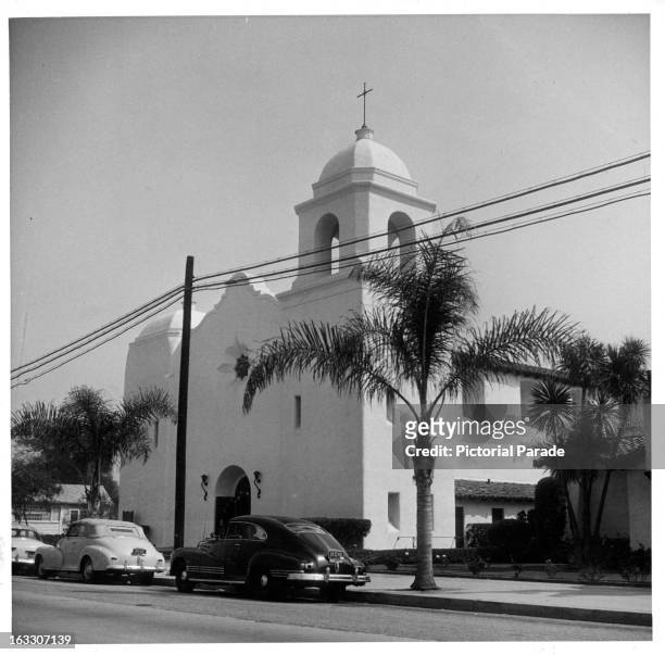 Street view of St Charles Church with cars parked curbside in front of the entrance, also in view, two young palm trees planted in front of the...