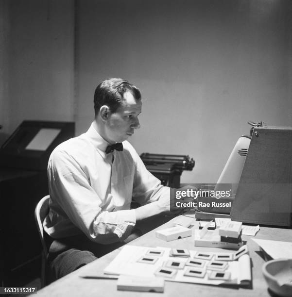 Man wearing a white shirt with a bow tie, sitting at a slide viewer, beside which are mounted 35mm transparencies, United States, circa 1950.