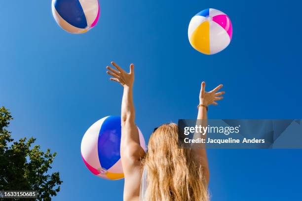 fun in the summer sun, a girl throws beach balls in the air under cloudless blue sky - beach ball stock pictures, royalty-free photos & images