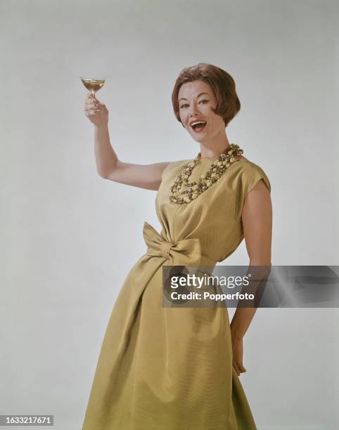 Posed studio portrait of a female fashion model wearing a light green dress with capped sleeves and bow waist and large beaded necklace, she raises a...