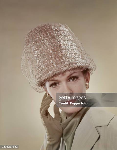 Posed studio portrait of a female fashion model wearing a sophisticated straw 'flowerpot' high hat, matching light grey coat, green neckscarf and...