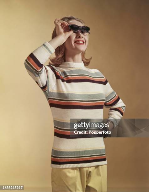 Posed studio portrait of a female fashion model wearing a knitted jersey roll necked sweater with black, grey and red stripes, she holds a pair of...