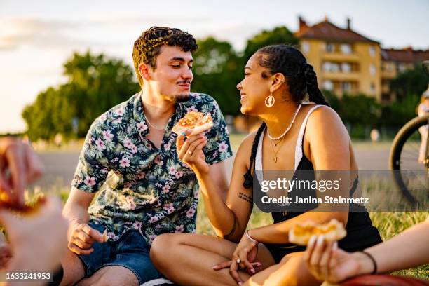 young couple look at each other in love while sitting in a park eating pizza - couple picnic stock pictures, royalty-free photos & images
