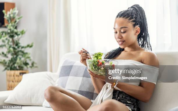 cute black girl eating yummy vegetable salad on sofa at home. slim african american teen maintaining weight by meatless food. - vegetarian food stock pictures, royalty-free photos & images