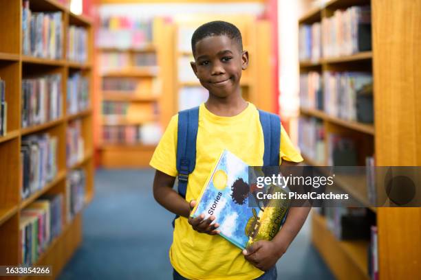 portrait boy with rucksack holding reading book standing in library between bookshelves - picture book stock pictures, royalty-free photos & images