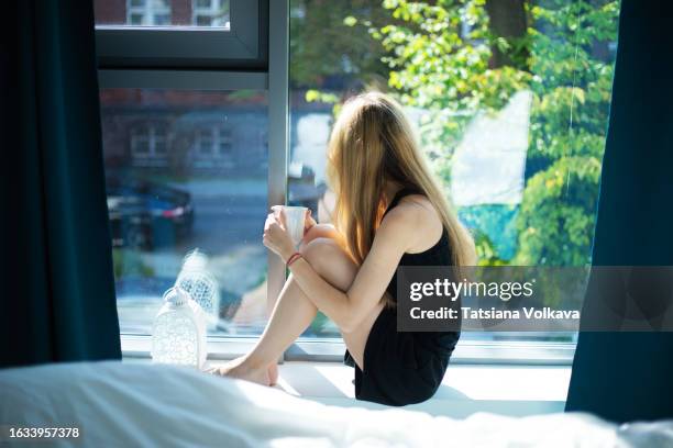 lovely teenage girl with long blond hair sits on window sill in bedroom looking at street - skinny teen stock pictures, royalty-free photos & images