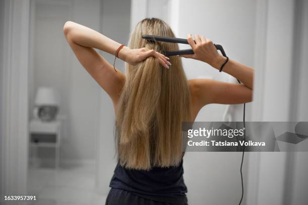 rear view of slim teenage girl with long blonde hair standing in corridor and using hair straightener - flat iron stock pictures, royalty-free photos & images