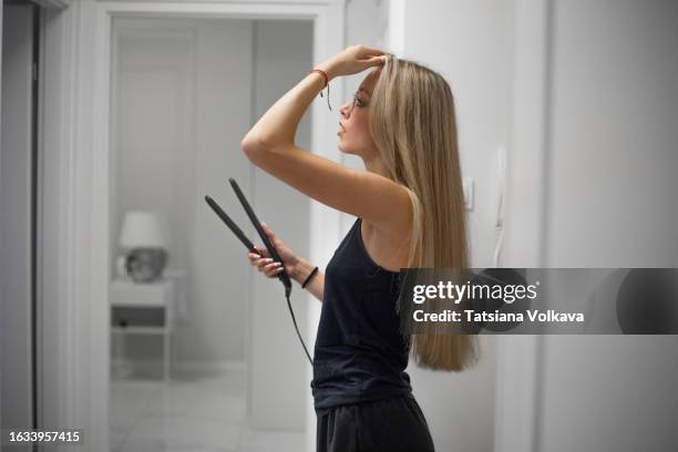 side view of attractive teenage girl with long blonde hair using hair straightener and looking in mirror - skinny girl stock pictures, royalty-free photos & images