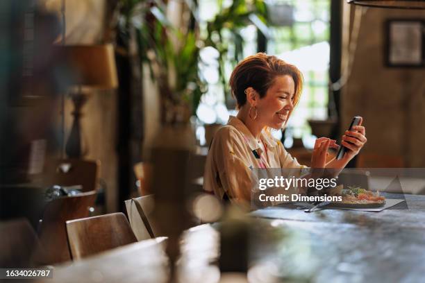 joven hispana feliz usando el teléfono mientras almuerza en un restaurante. - una mujer de mediana edad solamente fotografías e imágenes de stock