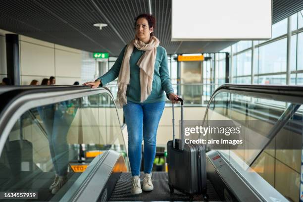 full length portrait of a beautiful woman standing on the stairs of the airport with her suitcase to the side, front view - woman flying scarf stock pictures, royalty-free photos & images