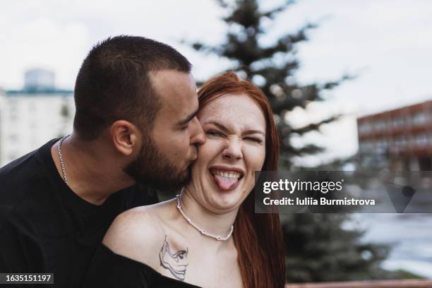 good-looking young man kissing attractive redhead woman on cheek while woman grimacing in discontent - couple tongue kissing stock-fotos und bilder