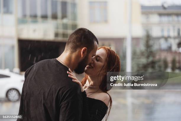 affectionate young couple stand on street kissing passionately unaware of warm summer rain falling - rain kiss stockfoto's en -beelden