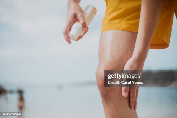 a male spritzes his leg with spray sun screen at beach - sonnenschutz stock-fotos und bilder