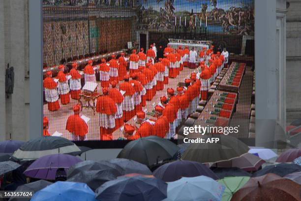 Faithful, holding umbrellas, look at a giant screen showing the start of the papal election conclave on St Peter's square on March 12, 2013 at the...