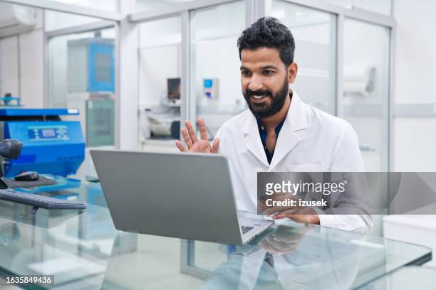 smiling male scientist waving while doing video call through laptop at desk in laboratory - clinical laboratory stock pictures, royalty-free photos & images