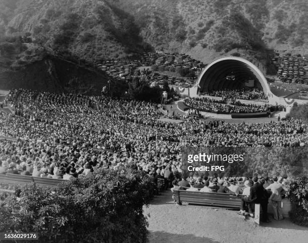View from the back of the amphitheatre as crowds of people watch a performance, with a large Stars-and-Stripes flag hanging on the stage of the...