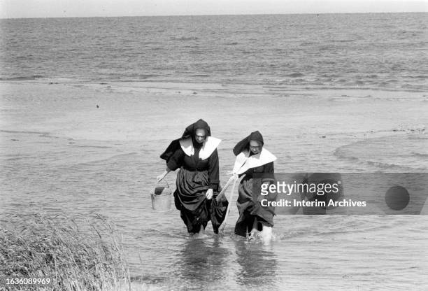 Pair of nuns hold the hem of their tunic as they wade along the beach while on the hunt for clams , Long Island, New York, 1957. One holds a bucket...