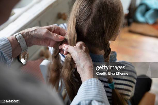 over the shoulder view of grandmother with arthritic hands, braiding a little girl's brown hair in a domestic setting. - tressé photos et images de collection