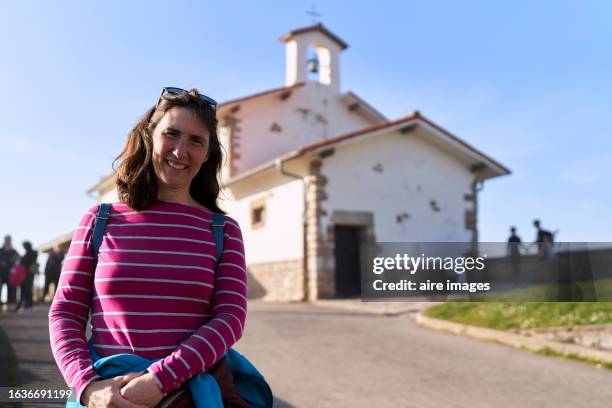 frontal portrait of a tourist woman with a backpack standing smiling looking towards the camera in the town of zumaia with a church in the background. - human face frontal stock pictures, royalty-free photos & images