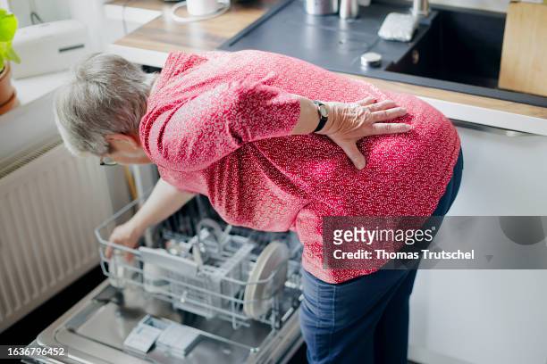 Symbolic photo on the subject of back pain when doing housework. An old woman loads a dishwasher with her back bent, holding her aching back on...