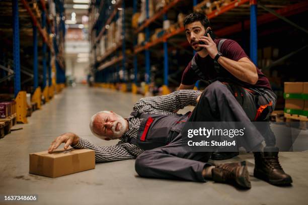 a painful manual worker lying down injured at the warehouse while his colleague is helping him. - trabajador manual stock pictures, royalty-free photos & images